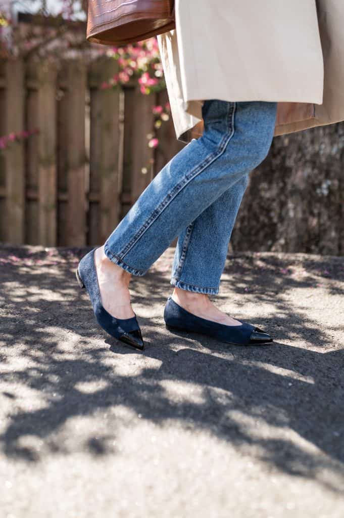 woman wearing a trench coat, ankle length blue jeans and comfortable flats in blue suede with a black patent cap toe