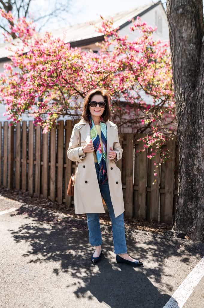 brunette woman wearing a trench coat with a colorful scarf, jeans, and comfortable flats that are blue suede in front of a blooming spring tree