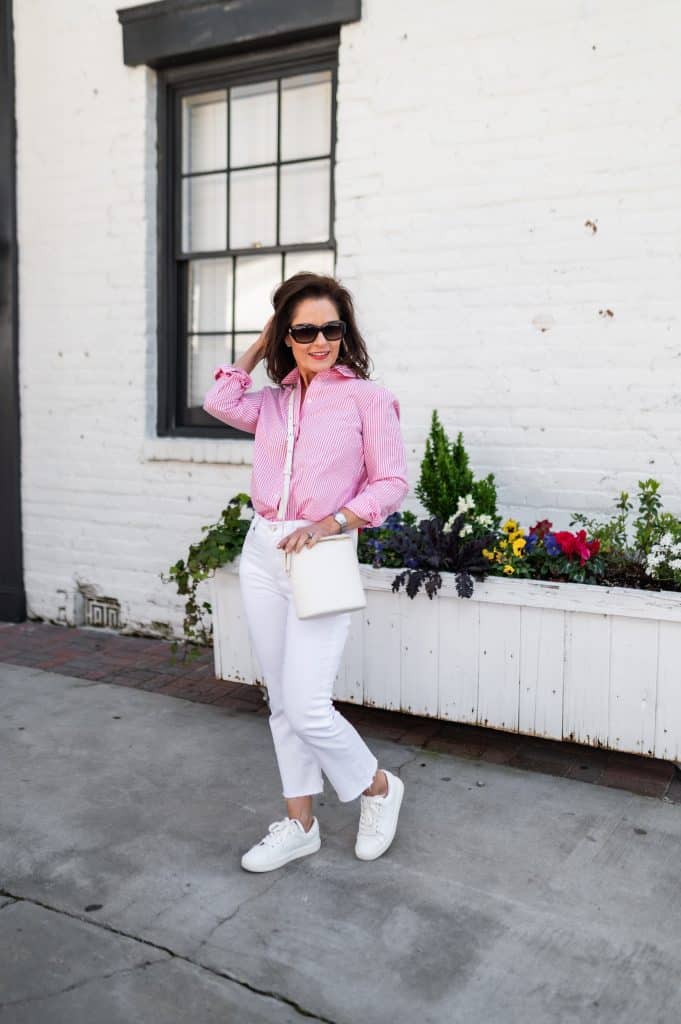 woman standing in front of a white brick wall and flower bed wearing a pink and white striped shirt, white jeans, and white sneakers, spring wardrobe classics
