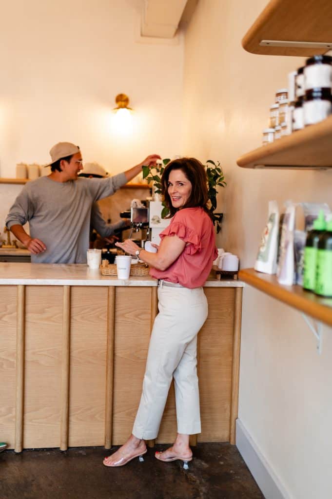 brunette woman standing at a counter getting a coffee wearing a pink top, khaki pants, and kitten heel comfortable heeled sandals