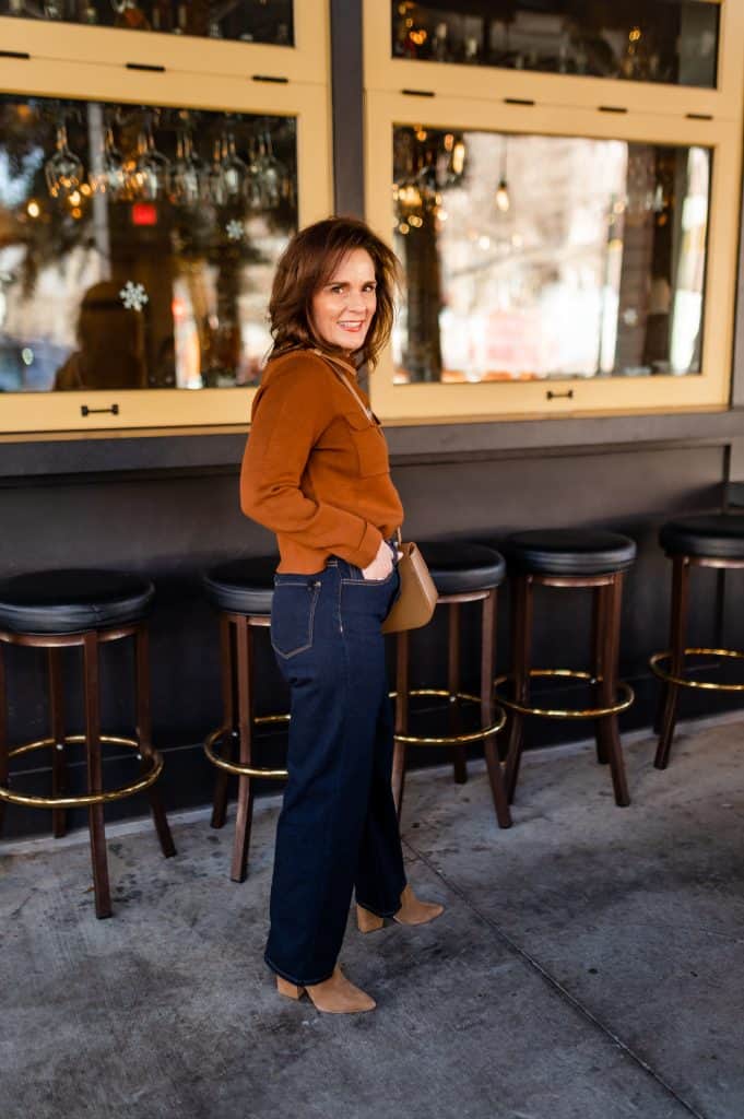 brunette woman wearing a brown sweater, wide leg jeans, and booties standing in front of a bar at a restaurant