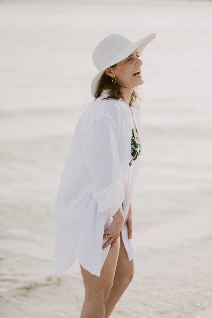 woman at the beach wearing a hat, bathing suite, and white bathing suit cover up, travel with a hat is a must