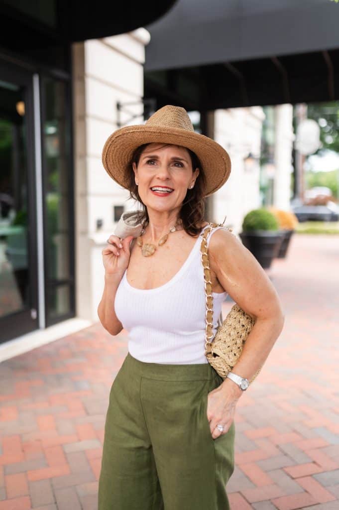 brunette woman wearing a white tank top, green pants and a straw hat, confidently showing her upper arms