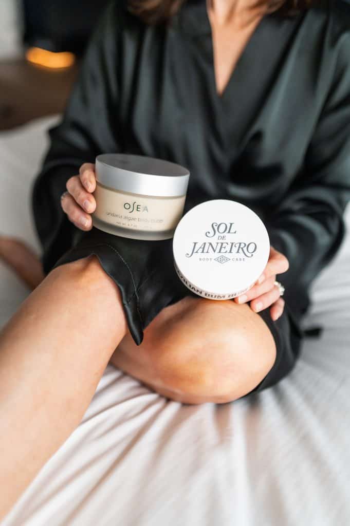 woman wearing a black robe holding a jar of body butter and  a jar of body lotion