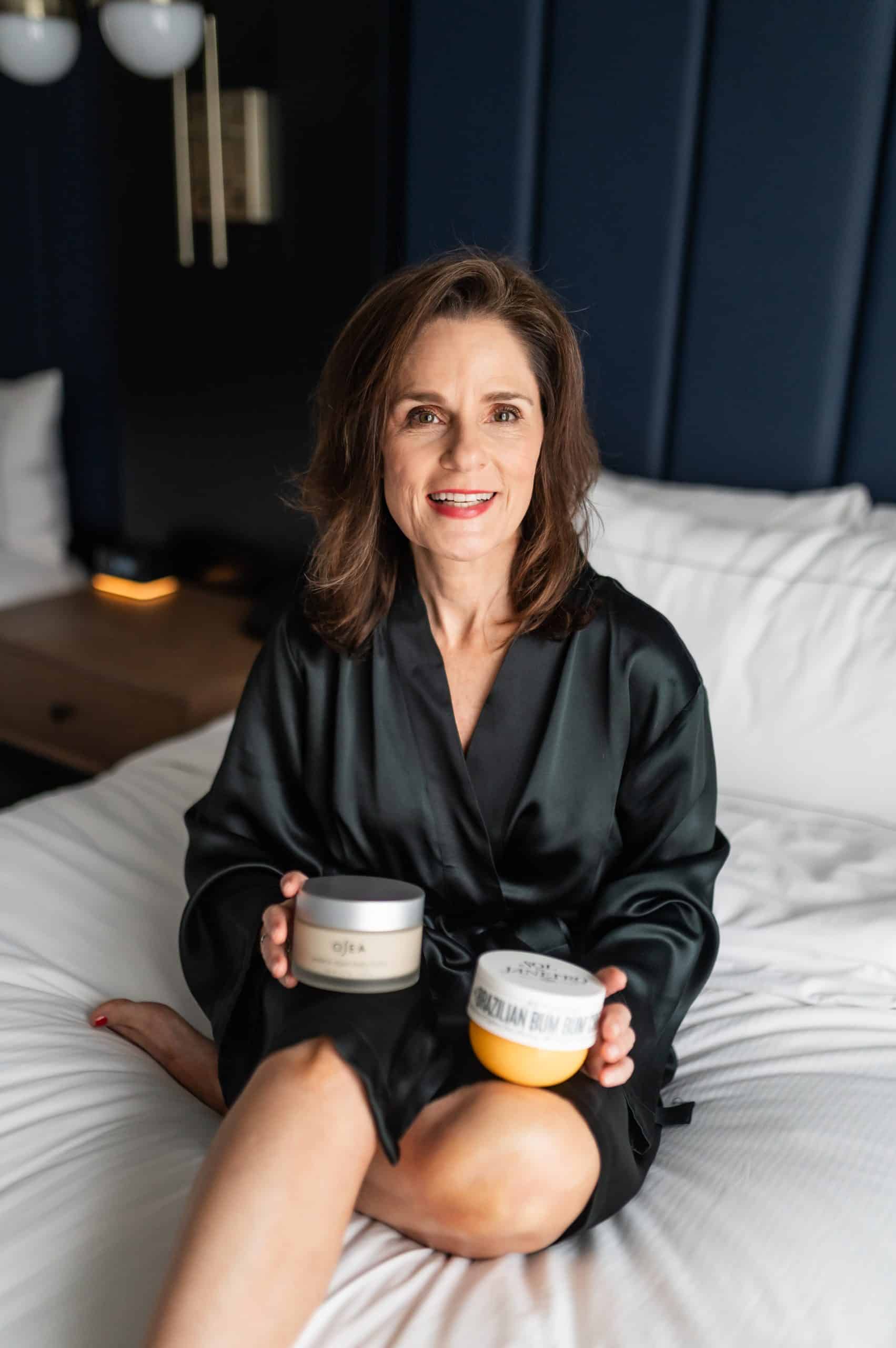 midlife woman sitting on a bed wearing a black silk robe with a jar of body butter in one hand and body lotion in the other