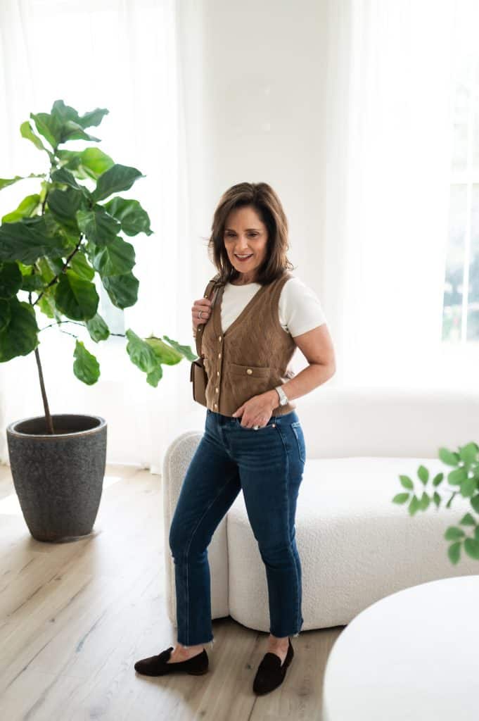 brunette woman wearing classic comfy loafers with jeans and a sweater vest standing in front of a white sofa