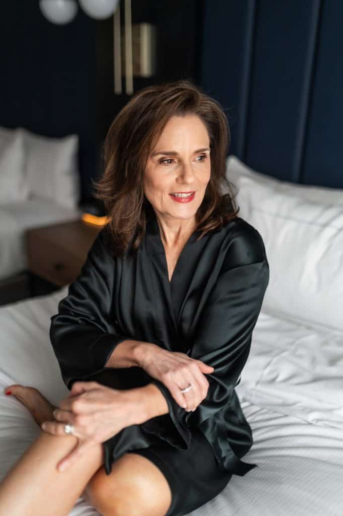 breunette woman sitting in the middle of a bed with white linens on it wearing a black silk robe