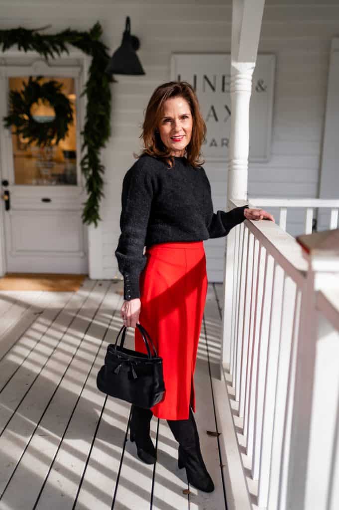 petite brunette woman wearing a red maxi skirt with a black sweater tucked into the waist and knee boots, the perfect skirt lengths of midi skirt for petites