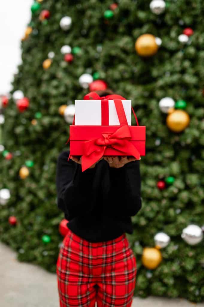 woman standing in front of a large Christmas tree holding red and white packages in front of her face