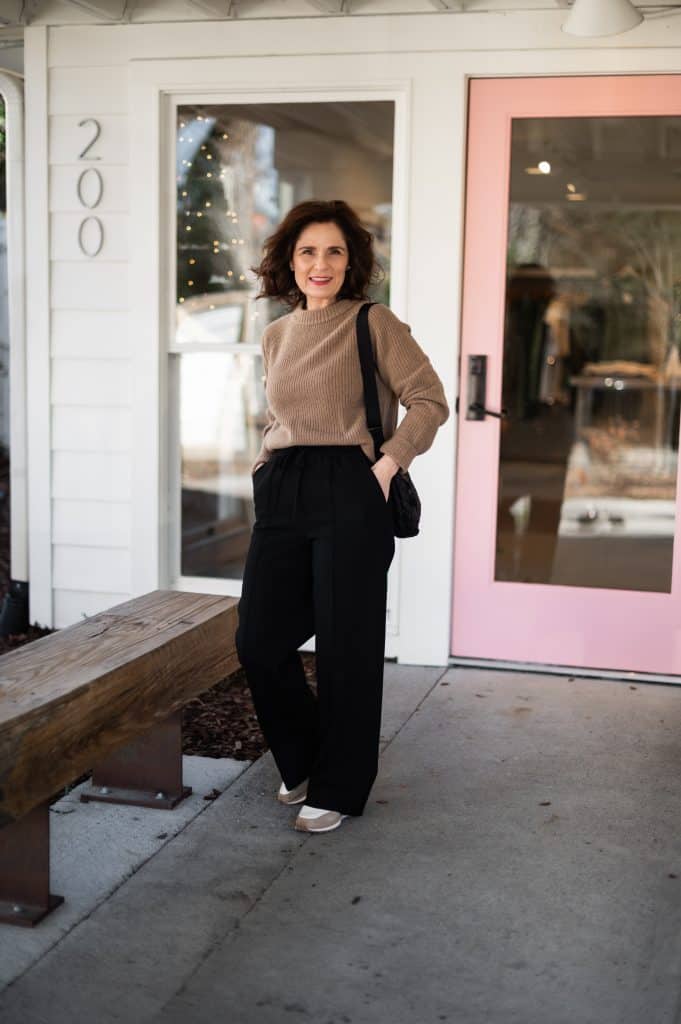 Sezane Bruce trousers styled on a petite woman wearing black wid-leg trousers with a neutral sweater and sneakers.